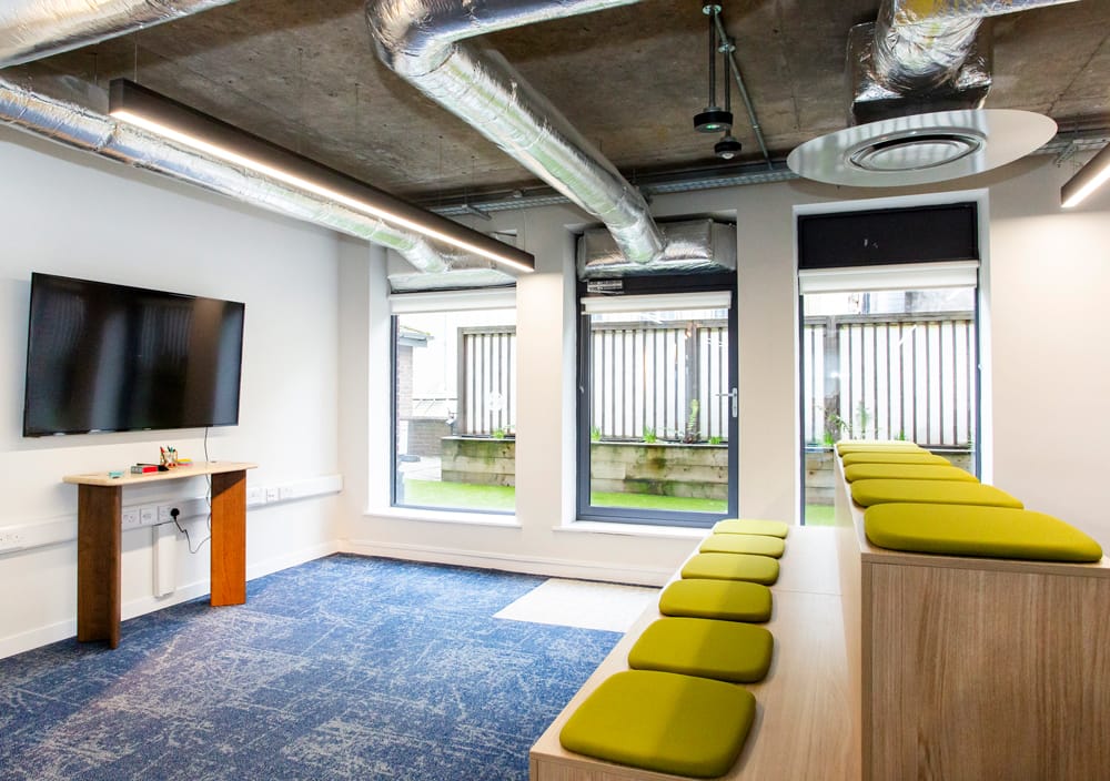 Office collaboration space with green cushioned seating, TV screen, and views to a courtyard