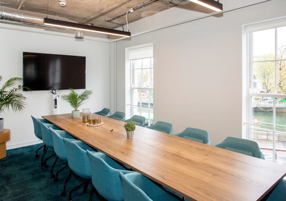 Boardroom-style meeting room with a long wooden table, teal chairs, and TV screen